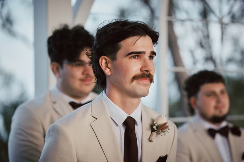 Joel, the groom, stands at the ceremony stage at Sandstone Point Hotel — The Pavilion, wearing a beige suit with a boutonniere, accompanied by two groomsmen in similar attire.