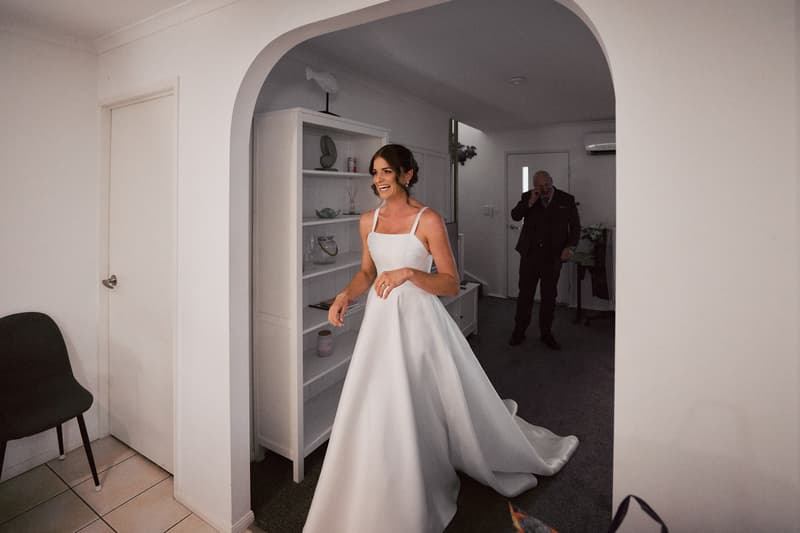 The bride Ashleigh in her wedding gown stands smiling inside a room at Sandstone Point Hotel, while an older man, likely the father of the bride, stands in the background near a door.