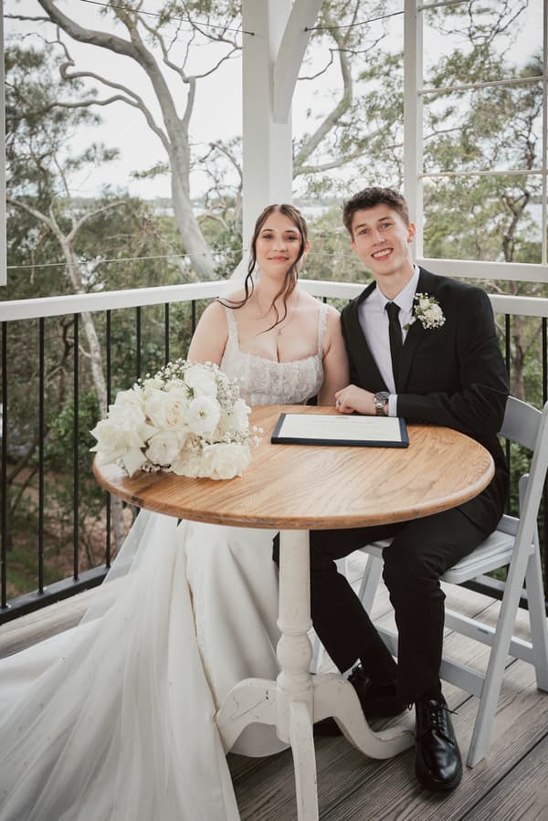 The bride Terion and groom Tyler sit at a round wooden table on a balcony at Sandstone Point Hotel — The Pavilion, with Terion's bouquet and a document on the table.