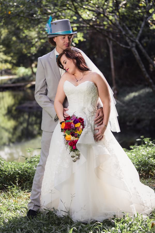 Bride Holly in a white wedding gown holding a colorful bouquet leans against groom Paul dressed in a light grey suit and a grey hat with a blue feather, standing outdoors near a pond at Kwila Lodge.