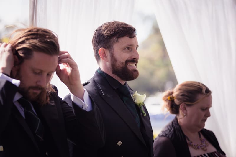 The groom and two guests stand near white draped fabric at the Toowong Rowing Club by the Brisbane River's edge during the ceremony stage.