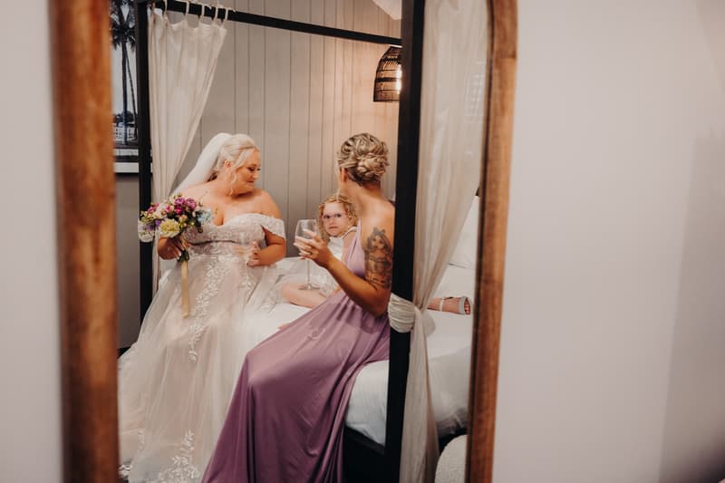 The bride Chantelle in her wedding gown holding a bouquet and a glass, sitting on a bed with a young girl and a bridesmaid in a lavender dress holding a glass, inside a room at Sandstone Point Hotel.