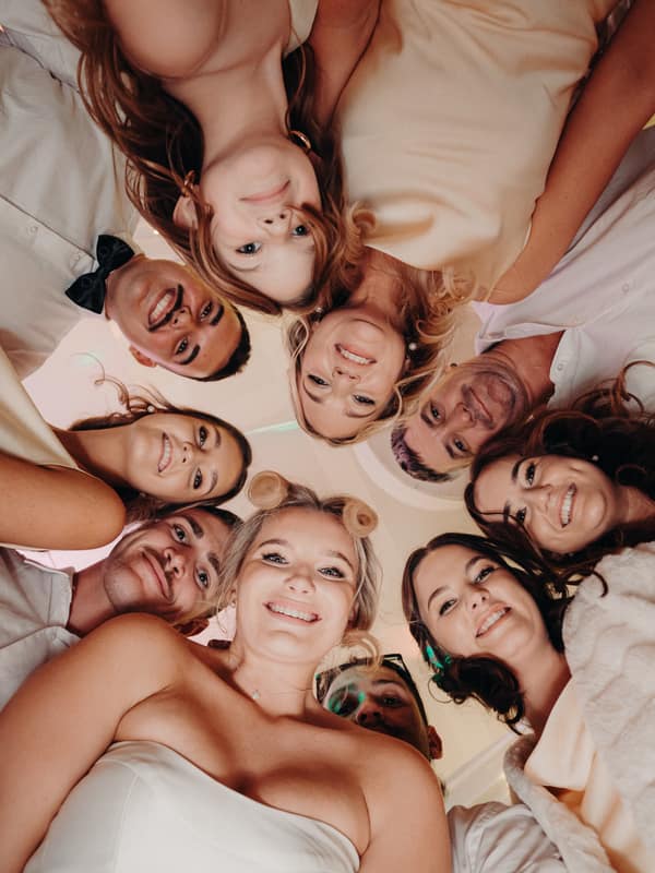 Libby the bride and Kyle the groom with their wedding party form a circle looking down at the camera inside The Tides — The Pandanus Room during the reception.