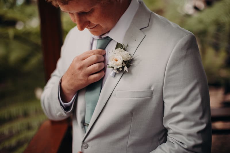 The groom Brodie adjusts his tie wearing a light grey suit with a white rose boutonniere at Sandstone Point Hotel.