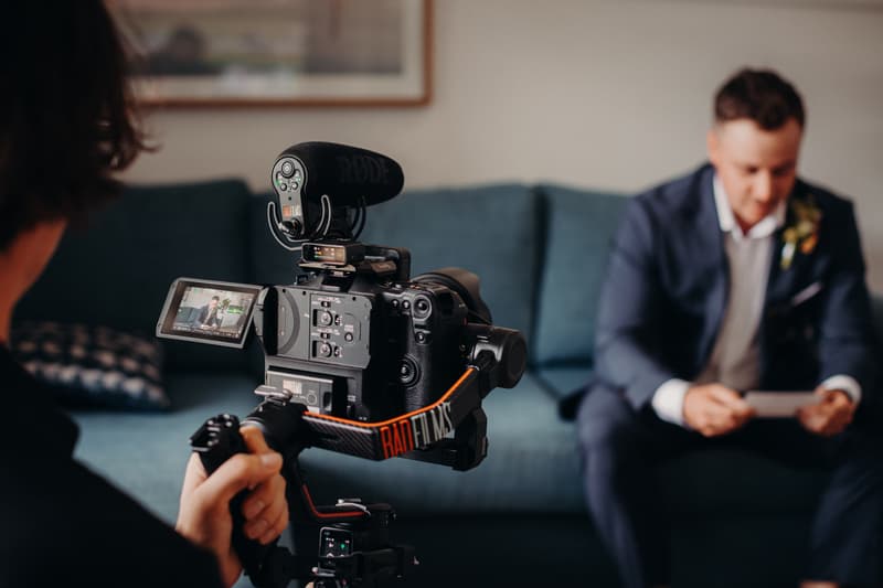 A videographer films Brandon the groom sitting on a blue couch at Sandstone Point Hotel, holding a card and wearing a dark suit with a boutonniere.