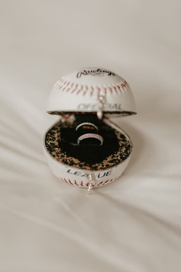 Two wedding rings displayed inside an open baseball-shaped ring box on a white fabric surface.
