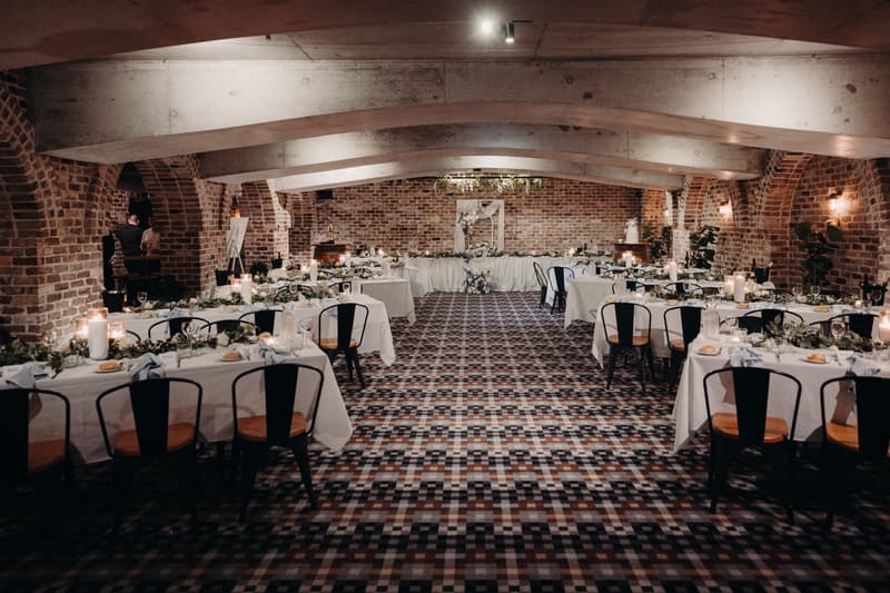 Reception tables set with white tablecloths, candles, greenery, and place settings in the Cellar at Sandstone Point Hotel, featuring exposed brick walls and arched ceilings.