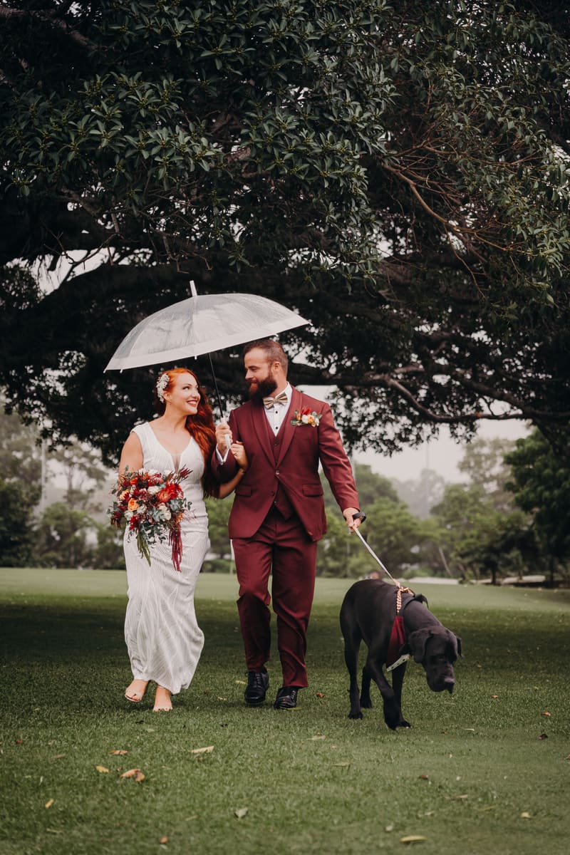 Red and Jayden walk together on grass under a large tree, The Pavilion, with Red holding a bouquet and Jayden holding a transparent umbrella and a black dog on a leash. Wedding family photos by She Said Yes.