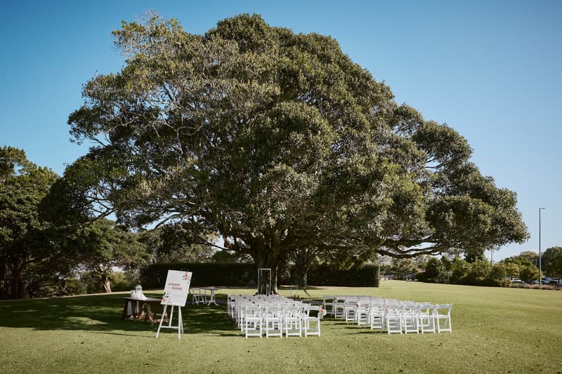 Ceremony setup at Sandstone Point Hotel — The Pavilion with white chairs arranged in rows facing a large tree, a sign with the names Jheng and Dane, and a small table nearby on a grassy lawn under a clear blue sky.