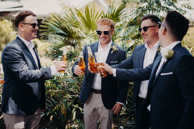 Four groomsmen wearing navy blazers, white shirts, and sunglasses toast with beer bottles outdoors at Sandstone Point Hotel.