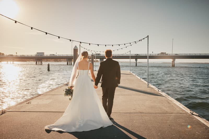 The bride Ashleigh in a white wedding gown and veil holding a bouquet and the groom James in a black suit walk hand in hand along a pier at Sandstone Point Hotel with water and a bridge in the background during sunset.
