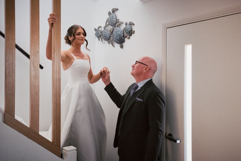 The bride Ashleigh stands on a staircase holding hands with an older man, likely her father, inside a building at Sandstone Point Hotel.