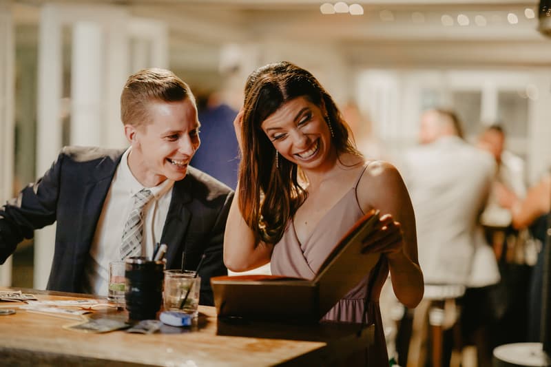 A man in a suit and tie and a woman in a sleeveless dress are seated at a wooden table with drinks, looking at a book or album together indoors with other guests blurred in the background.