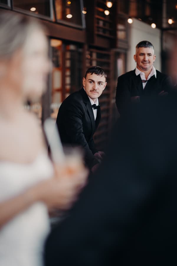 Kyle in a tuxedo seated indoors at The Tides — The Pandanus Room reception, looking towards the bride Libby who is blurred in the foreground holding a drink; another man stands behind Kyle.
