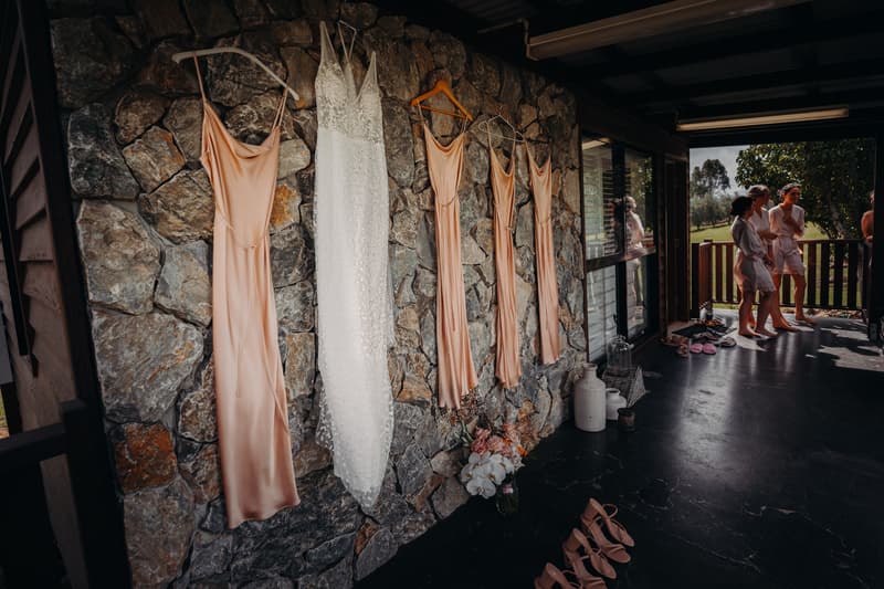 The bride's white wedding dress and four bridesmaids' peach dresses hang on a stone wall at Preston Peak Winery Functions Centre, with bridesmaids in robes standing on the porch in the background.