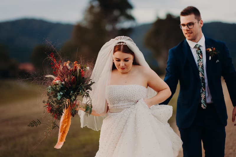 The bride Lilly in a strapless white wedding gown and veil holds a bouquet of flowers while walking outdoors with the groom Connor, who is wearing a dark blue suit and floral tie, at Yabbaloumba Retreat.