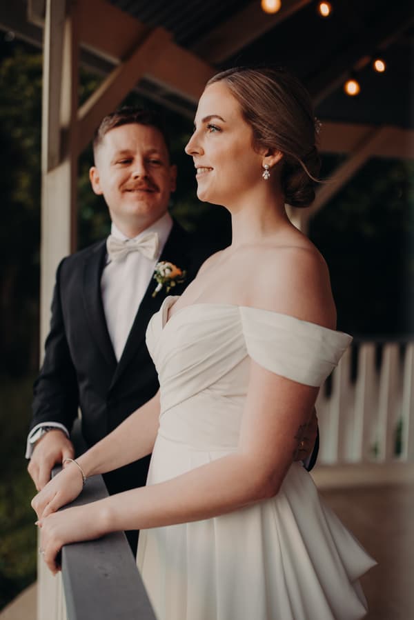 Courtney and Liam stand together on a veranda at Tiffany's Maleny, with Courtney in an off-shoulder wedding gown and Liam in a black suit with a white bow tie and boutonniere.