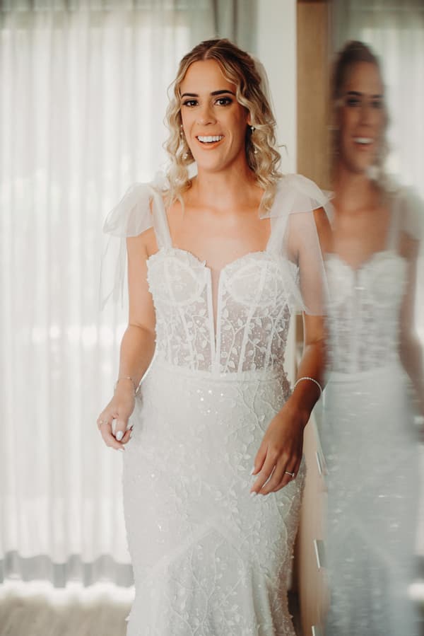 The bride Krystal stands indoors near a reflective surface wearing a white lace wedding gown with sheer shoulder straps and intricate floral detailing.