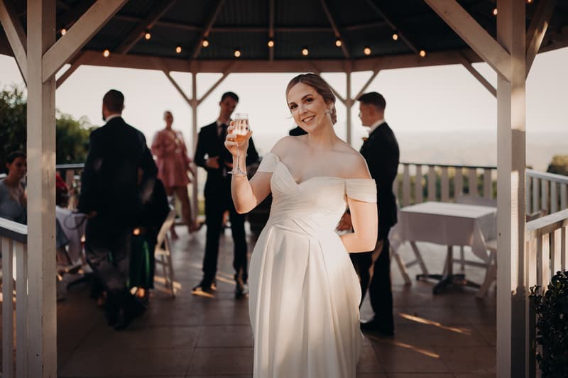 The bride Courtney in a white off-shoulder gown holds a glass of champagne and smiles at the camera on the reception stage at Tiffany's Maleny — Quartyard, with guests mingling in the background under a pavilion.