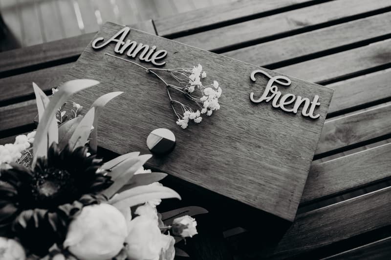 A wooden board with the names Annie and Trent placed on a wooden slatted surface, accompanied by small flowers and a bouquet partially visible in the foreground at Ocean View Estates.