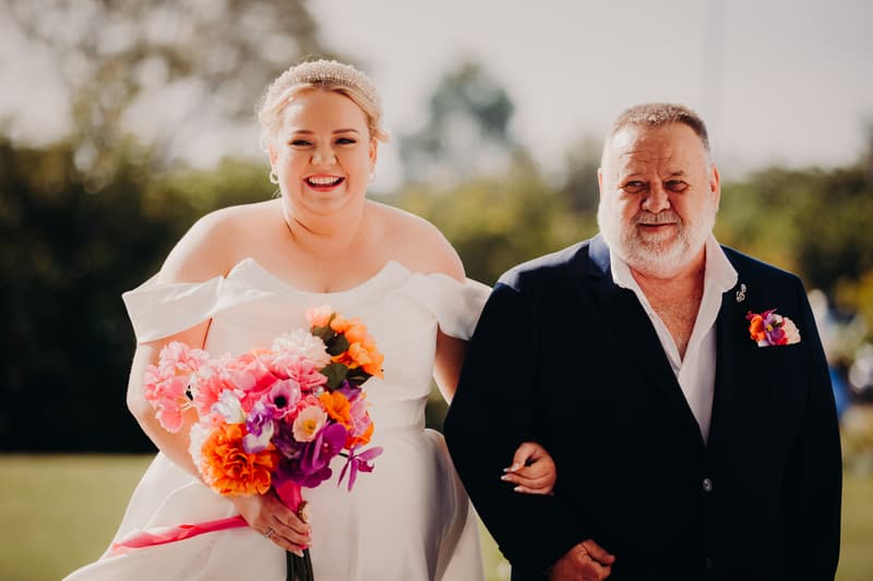 The bride Jacquelyne, holding a colorful bouquet, is walking arm-in-arm with an older man, likely her father, outdoors at Sandstone Point Hotel — Pavilion.