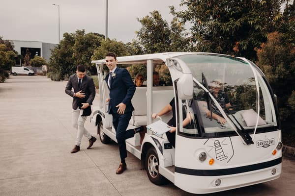 The groom Cameron in a navy suit steps off a white golf cart with a boutonniere pinned to his lapel at Sandstone Point Hotel — Pavilion, accompanied by a man in a dark blazer and light pants, with other seated guests visible inside the cart.