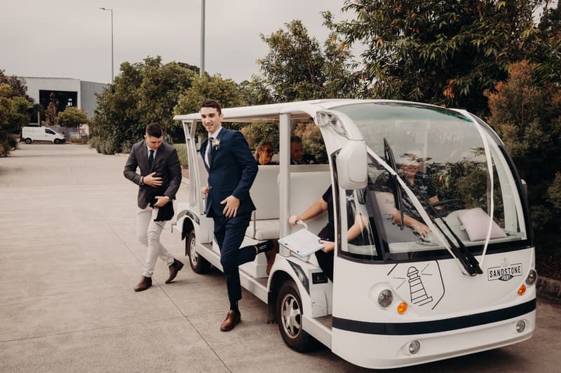 The groom Cameron in a navy suit steps off a white golf cart with a boutonniere pinned to his lapel at Sandstone Point Hotel — Pavilion, accompanied by a man in a dark blazer and light pants, with other seated guests visible inside the cart.