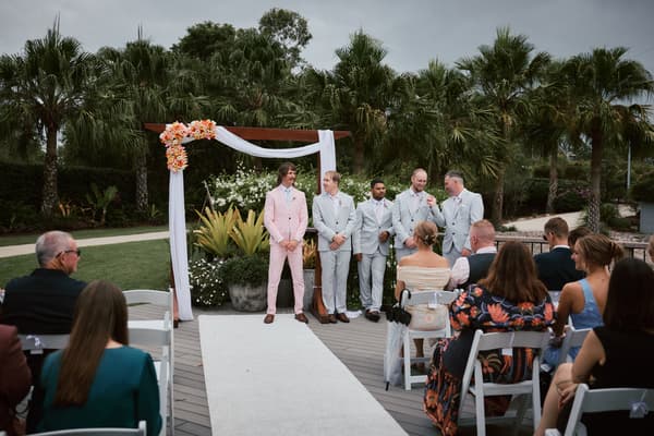 Jake and his groomsmen stand at the ceremony arch decorated with white fabric and orange flowers at Eatons Hill Hotel — Lakeside, while guests are seated watching.