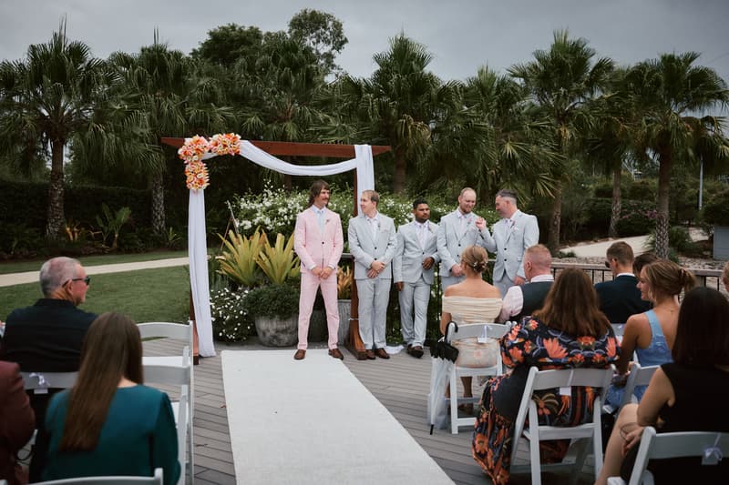 Jake and his groomsmen stand at the ceremony arch decorated with white fabric and orange flowers at Eatons Hill Hotel — Lakeside, while guests are seated watching.