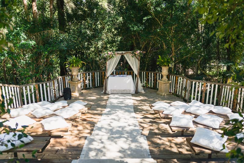 Empty outdoor ceremony setup at Kwila Lodge with a white aisle runner leading to a small altar draped with white fabric and floral arrangements, surrounded by wooden benches with white cushions.