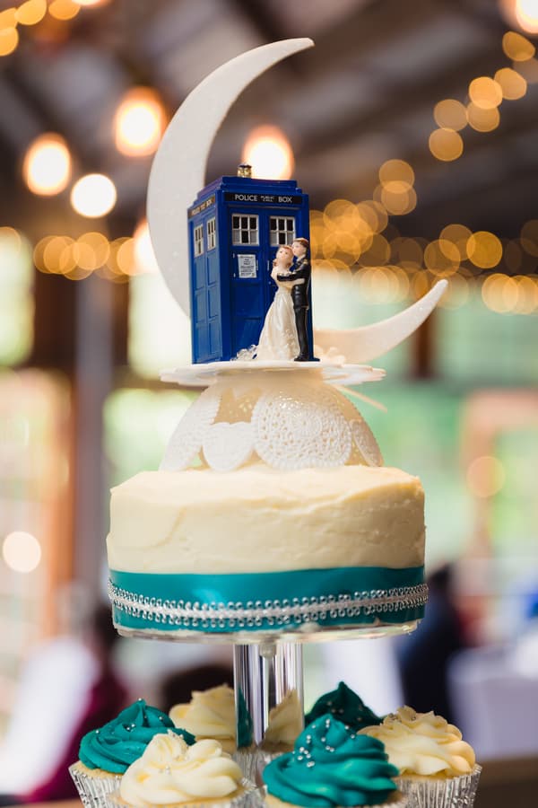 Wedding cake with a topper featuring a bride and groom figurine standing next to a blue police box and a crescent moon, displayed at Kwila Lodge reception.