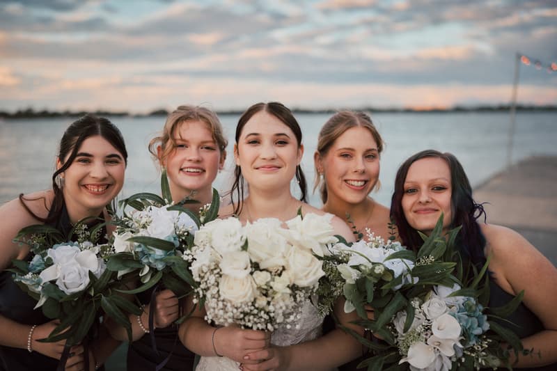 The bride Terion poses with four bridesmaids holding white floral bouquets outdoors near water at Sandstone Point Hotelallo during the couple portraits stage.