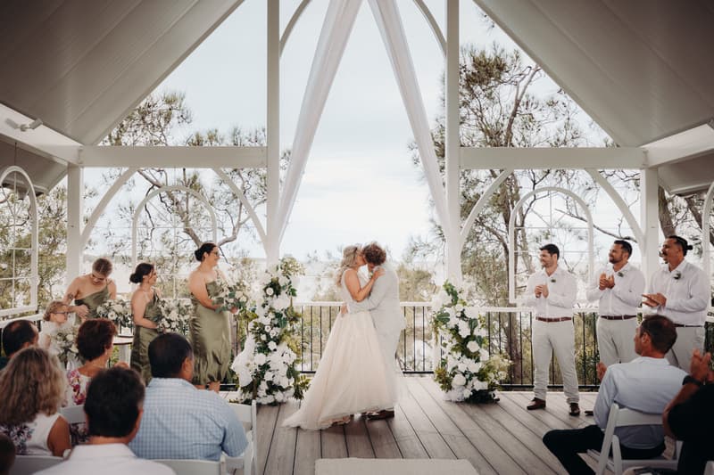 Bride Chloe and groom Brodie kiss at the altar during their wedding ceremony at Sandstone Point Hotel — Pavilion, surrounded by bridesmaids in olive green dresses, groomsmen in white shirts and light pants, and seated guests.