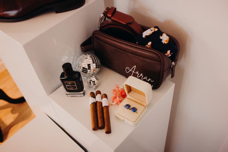 Groom's leather toiletry bag labeled 'Arran' with a pair of blue cufflinks in an open box, three cigars, a bottle of cologne, two small disco ball ornaments, and a pair of patterned socks on a white shelf.