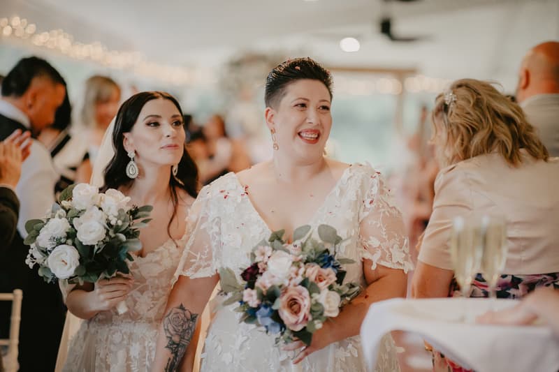 Two brides in white lace wedding dresses holding bouquets stand together indoors with guests in the background.