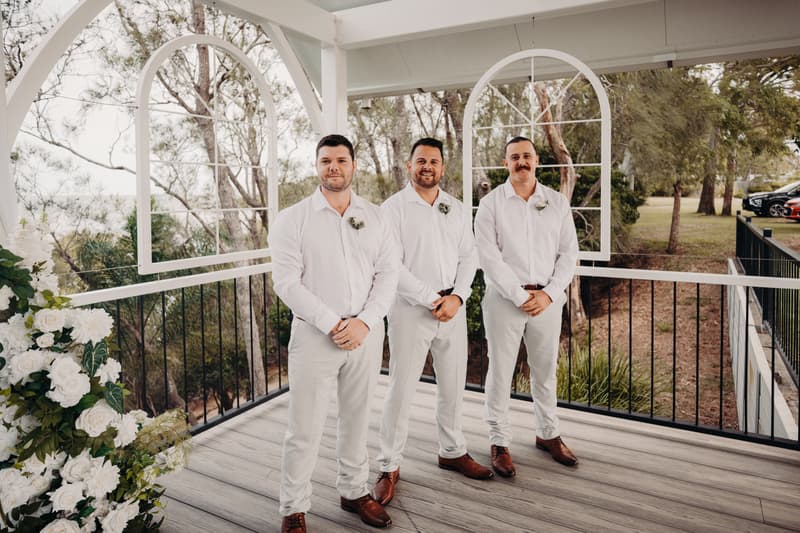 Three groomsmen stand side by side on the Pavilion deck at Sandstone Point Hotel, dressed in white shirts and light-colored pants with brown shoes, with floral boutonnieres pinned to their shirts and white floral arrangements visible to the left.