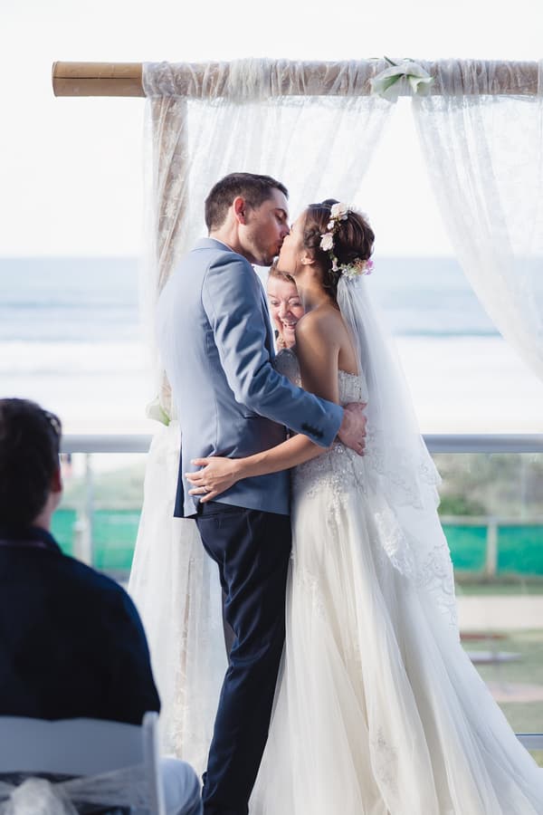 Bride Wing and groom Jason share a kiss at the altar during their ceremony at Bilinga Beach Weddings — The Terrace, with an officiant standing behind them and a guest seated in the foreground.