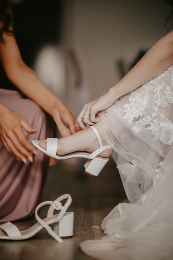 A person in a mauve dress helps the bride put on a white heeled sandal, with the bride wearing a lace wedding gown.