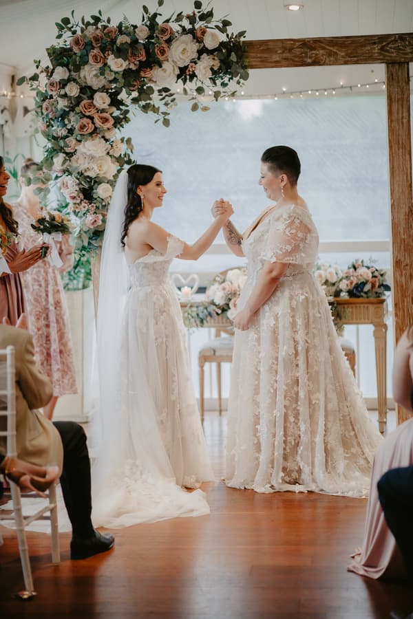 Two brides in lace wedding dresses hold hands facing each other under a floral arch during a wedding ceremony, with guests seated nearby.