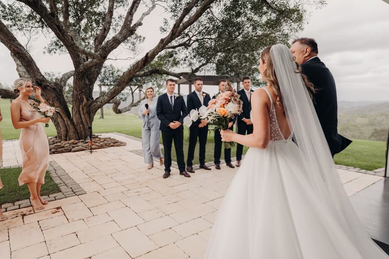The bride Chelsea, accompanied by an older man, stands holding a bouquet at the Preston Peak Winery Functions Centre ceremony stage, facing the groom Brent and his groomsmen, with a bridesmaid holding flowers standing to the side.
