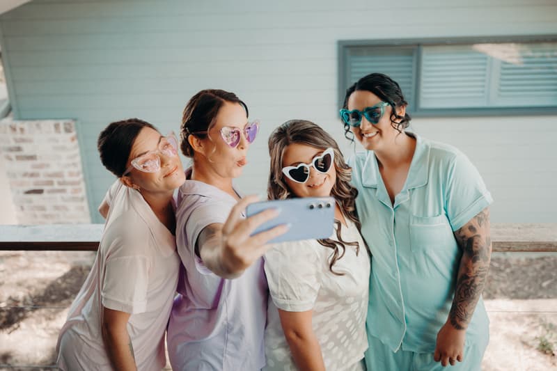 Four bridesmaids wearing colorful heart-shaped sunglasses take a selfie together at Sandstone Point Hotel — The Pavilion.