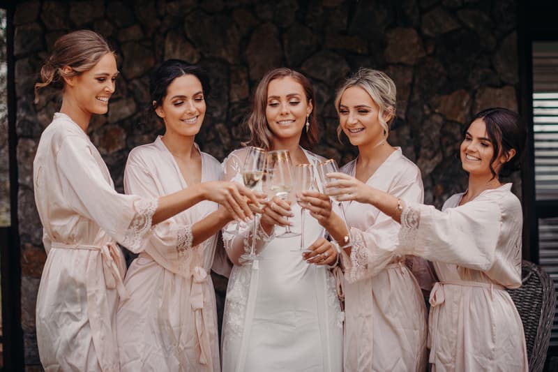The bride Chelsea in a white dress and four bridesmaids in matching light pink robes toast with champagne glasses at Preston Peak Winery Functions Centre.