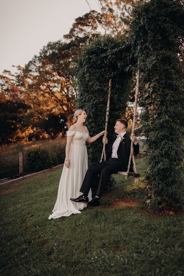 Courtney, the bride, stands beside Liam, the groom, who is seated on a wooden swing wrapped in greenery at Tiffany's Maleny outdoors during their couple portraits session.