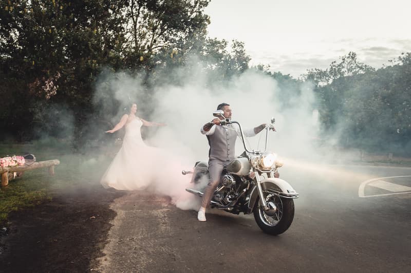 The groom rides a motorcycle creating smoke while the bride stands behind him in her wedding dress at Novotel Sunshine Coast Resort outdoors.