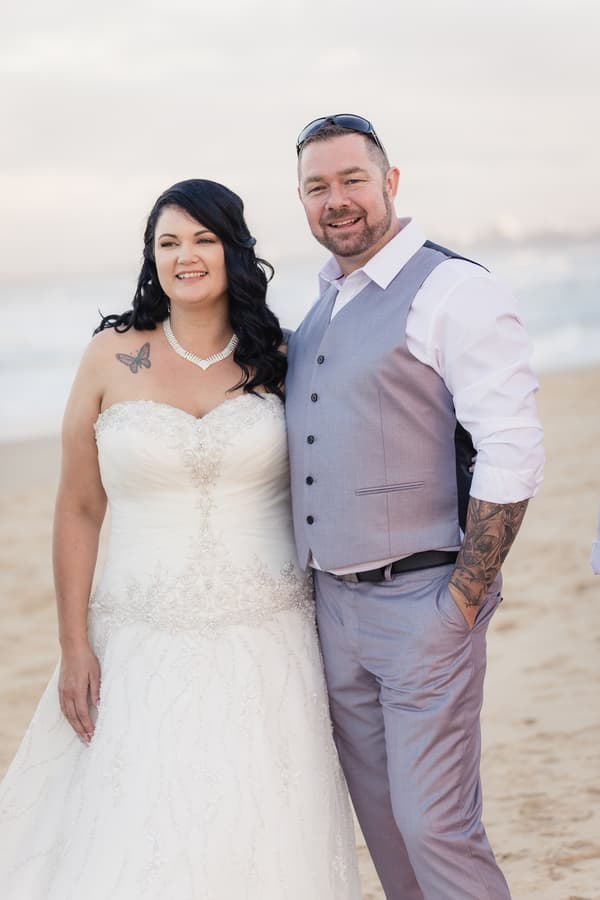 The bride Kelly and groom Shane pose together on a sandy beach with the ocean in the background.