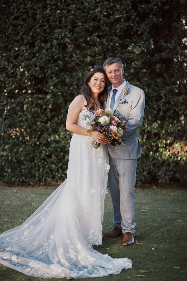 The bride Jheng and groom Dane stand together on grass in front of dense greenery at Sandstone Point Hotel. Jheng wears a white floral lace wedding gown and holds a bouquet of flowers. Dane wears a light grey suit with a blue tie and boutonniere.