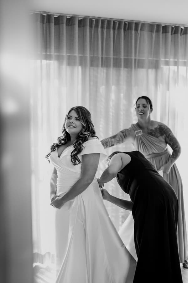 The bride Brooke stands in a white wedding gown while two attendants assist her with the dress at Sandstone Point Hotel — The Pavilion.
