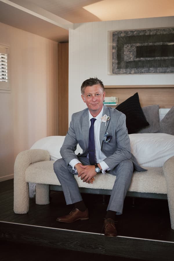 The groom Dane sits on a bench in a room at Sandstone Point Hotel, wearing a grey suit with a boutonniere and brown shoes.
