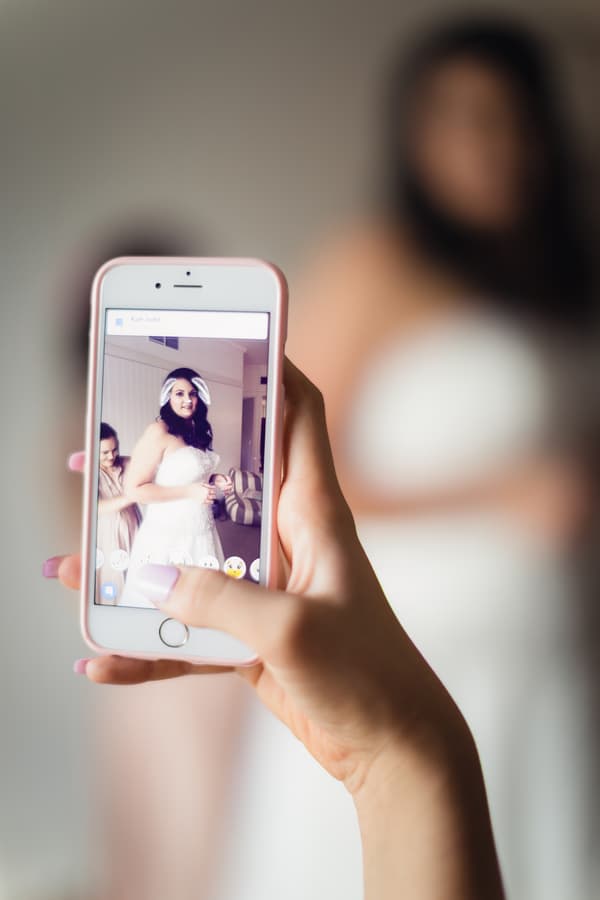 The bride Kelly is shown on a smartphone screen wearing her wedding dress with a digital bunny ears filter applied, while another person adjusts her dress in the background at Novotel Sunshine Coast Resort.