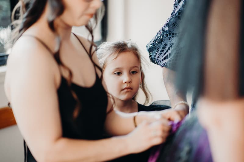 A young girl looks on as a woman in a black dress adjusts the attire of another person indoors.